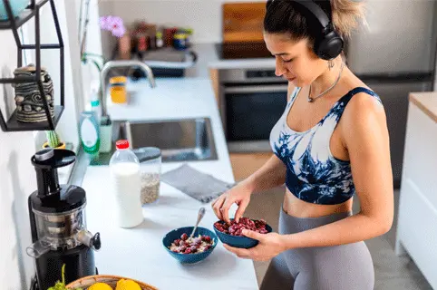 Eine sportliche Frau mit Kopfhörern bereitet in einer modernen Küche eine gesunde Mahlzeit vor. Sie hält eine Schüssel mit Obst und Joghurt in der Hand, während sie eine weitere Schüssel auf der Arbeitsfläche hat. Im Hintergrund sind Küchenutensilien und eine Obstschale sichtbar.