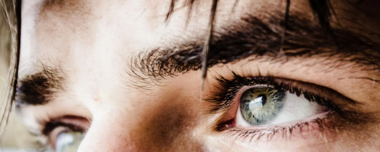 Close-up of a person's eyes, showcasing distinct, bright green irises surrounded by dark lashes and eyebrows. The focus is on the expressive gaze, suggesting curiosity or contemplation. The lighting highlights the details of the eyes and skin texture.